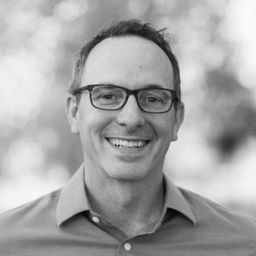 Robert Jolly smiling, wearing glasses and a collared shirt, outdoors with a softly blurred background.